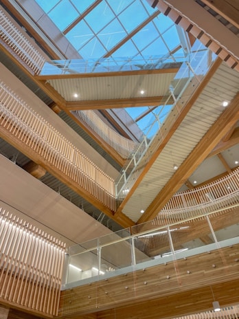 A modern architectural interior featuring a series of wooden beams and glass railings. The skylight ceiling allows natural light to flood the space, highlighting the clean lines and geometric design elements. The wood tones add warmth and contrast with the sleekness of the glass and metal.