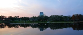 A serene landscape with a calm lake reflecting trees and a building in the background. The sky has soft hues of blue and pink as the sun sets, creating a peaceful atmosphere.