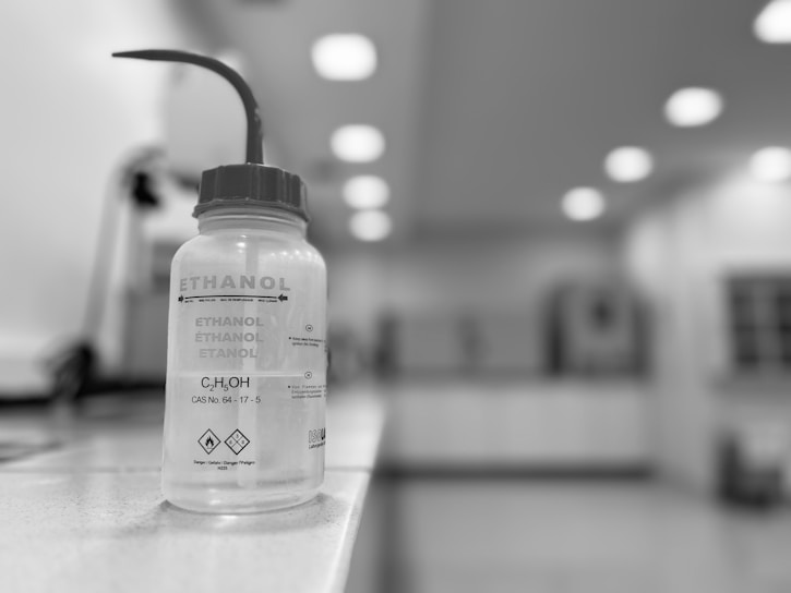 A plastic bottle labeled 'ETHANOL' with a science formula and warning symbols is placed on a laboratory counter. The bottle has a dispensing nozzle, and the background consists of laboratory equipment and a well-lit room with several ceiling lights.