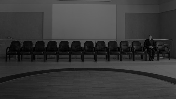 A row of empty black chairs lines a wall in an indoor setting, with a single person seated near the far end. The atmosphere is quiet and somber, with a stark, minimalist design comprising muted tones and minimal decor.