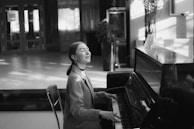 A person is seated at a piano, playing music with a peaceful expression. The setting appears to be indoors, with soft lighting and shadow patterns on the floor and walls. The background has a modern interior with plants and furniture.