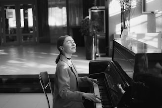 A person is seated at a piano, playing music with a peaceful expression. The setting appears to be indoors, with soft lighting and shadow patterns on the floor and walls. The background has a modern interior with plants and furniture.