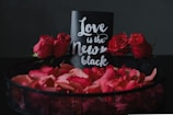 A decorative arrangement featuring a black card with white cursive text reading 'Love is the New Black', surrounded by vibrant red roses and rose petals in a dark basket. The backdrop is a dark, neutral color that accentuates the vivid red of the flowers and petals.