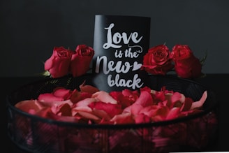 A decorative arrangement featuring a black card with white cursive text reading 'Love is the New Black', surrounded by vibrant red roses and rose petals in a dark basket. The backdrop is a dark, neutral color that accentuates the vivid red of the flowers and petals.