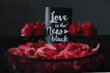 A decorative arrangement featuring a black card with white cursive text reading 'Love is the New Black', surrounded by vibrant red roses and rose petals in a dark basket. The backdrop is a dark, neutral color that accentuates the vivid red of the flowers and petals.