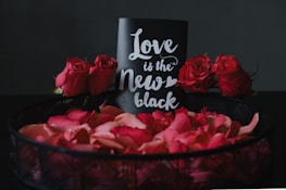 A decorative arrangement featuring a black card with white cursive text reading 'Love is the New Black', surrounded by vibrant red roses and rose petals in a dark basket. The backdrop is a dark, neutral color that accentuates the vivid red of the flowers and petals.