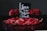 A decorative arrangement featuring a black card with white cursive text reading 'Love is the New Black', surrounded by vibrant red roses and rose petals in a dark basket. The backdrop is a dark, neutral color that accentuates the vivid red of the flowers and petals.