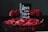 A decorative arrangement featuring a black card with white cursive text reading 'Love is the New Black', surrounded by vibrant red roses and rose petals in a dark basket. The backdrop is a dark, neutral color that accentuates the vivid red of the flowers and petals.
