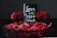 A decorative arrangement featuring a black card with white cursive text reading 'Love is the New Black', surrounded by vibrant red roses and rose petals in a dark basket. The backdrop is a dark, neutral color that accentuates the vivid red of the flowers and petals.