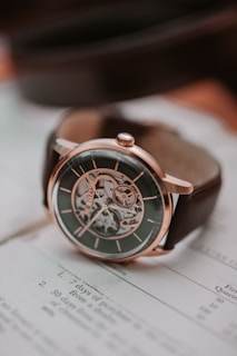 A sophisticated mechanical watch with a metallic case and visible inner workings, placed on top of an open book with text. The watch has a dark face, intricate gear design, and a brown leather strap.