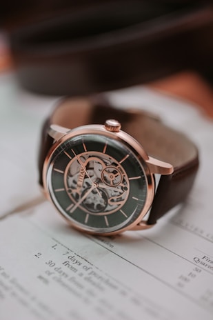 A sophisticated mechanical watch with a metallic case and visible inner workings, placed on top of an open book with text. The watch has a dark face, intricate gear design, and a brown leather strap.
