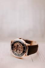 A close-up view of an elegant wristwatch with a black dial and a visible mechanical movement. The watch features a rose gold case and a dark brown leather strap. It is resting on a light-colored surface, which gives the picture a minimalist and sophisticated appearance.