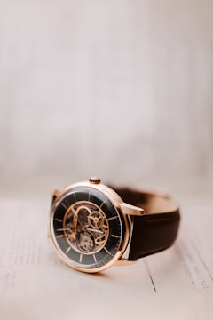 A close-up view of an elegant wristwatch with a black dial and a visible mechanical movement. The watch features a rose gold case and a dark brown leather strap. It is resting on a light-colored surface, which gives the picture a minimalist and sophisticated appearance.