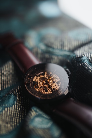 A close-up of a wristwatch with a brown leather strap resting on a textured fabric. The watch features an intricately designed skeleton dial with visible gears and mechanisms, and the word 'Fossil' is visible.