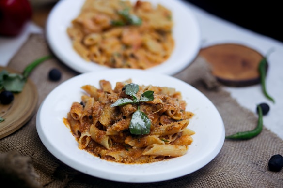 Two dishes of pasta served on white plates are present, one in the foreground with a rich tomato-based sauce and garnished with basil, and another slightly blurred in the background. The setting includes a rustic burlap cloth, green chili peppers, and a few black olives scattered around.