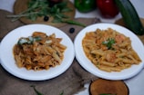 Two white plates of pasta dishes are placed on a textured brown fabric. The pasta dishes appear to be garnished with herbs and cheese. Surrounding the plates are fresh vegetables like green and red bell peppers, a zucchini, green chilies, and leafy greens. A wooden board sits in the background with some more leaves on it. The overall setting is rustic and seems to depict a cozy, home-cooked meal.