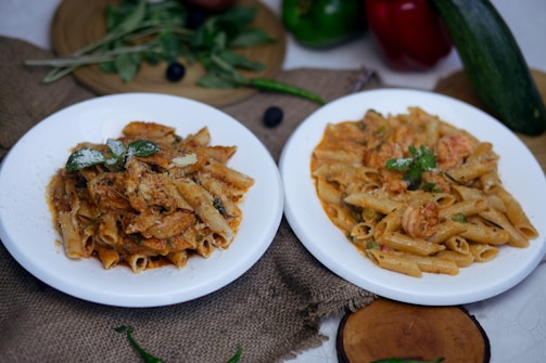 Two white plates of pasta dishes are placed on a textured brown fabric. The pasta dishes appear to be garnished with herbs and cheese. Surrounding the plates are fresh vegetables like green and red bell peppers, a zucchini, green chilies, and leafy greens. A wooden board sits in the background with some more leaves on it. The overall setting is rustic and seems to depict a cozy, home-cooked meal.