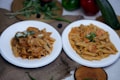 Two white plates of pasta dishes are placed on a textured brown fabric. The pasta dishes appear to be garnished with herbs and cheese. Surrounding the plates are fresh vegetables like green and red bell peppers, a zucchini, green chilies, and leafy greens. A wooden board sits in the background with some more leaves on it. The overall setting is rustic and seems to depict a cozy, home-cooked meal.