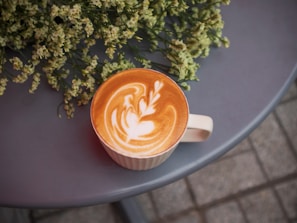 A cup of latte with intricate latte art sits on a gray round table. Beside it are delicate white flowers with green stems, creating a tranquil setting.