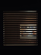A window with horizontal blinds partially covers a view of an illuminated interior space. Through the slats, a vibrant and colorful scene, possibly a mural or artwork, is visible, with hints of blues, oranges, and other colors.
