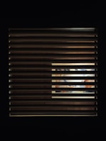 A window with horizontal blinds partially covers a view of an illuminated interior space. Through the slats, a vibrant and colorful scene, possibly a mural or artwork, is visible, with hints of blues, oranges, and other colors.