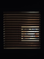 A window with horizontal blinds partially covers a view of an illuminated interior space. Through the slats, a vibrant and colorful scene, possibly a mural or artwork, is visible, with hints of blues, oranges, and other colors.