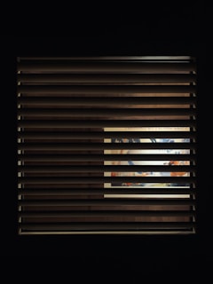 A window with horizontal blinds partially covers a view of an illuminated interior space. Through the slats, a vibrant and colorful scene, possibly a mural or artwork, is visible, with hints of blues, oranges, and other colors.