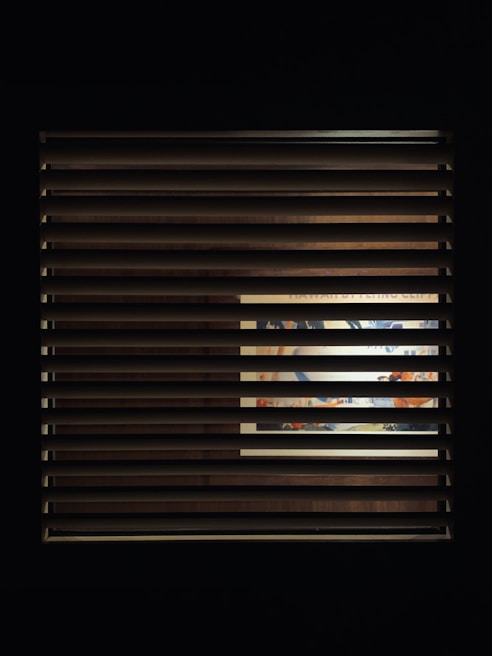 A window with horizontal blinds partially covers a view of an illuminated interior space. Through the slats, a vibrant and colorful scene, possibly a mural or artwork, is visible, with hints of blues, oranges, and other colors.