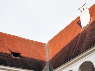 A section of a building with a steeply pitched red roof featuring a chimney and small window. The roof is divided by a strip of material, possibly for waterproofing. The bottom part of the image includes a segment of an arched structure in white.