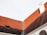 A section of a building with a steeply pitched red roof featuring a chimney and small window. The roof is divided by a strip of material, possibly for waterproofing. The bottom part of the image includes a segment of an arched structure in white.