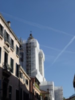 A street view of a historic building under renovation. The structure is wrapped in white scaffolding, with its dome prominently visible against the clear blue sky. The surrounding buildings have traditional architecture with narrow windows and shuttered balconies, adding to the historical ambiance.