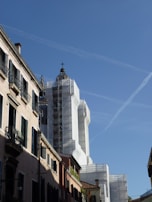 A street view of a historic building under renovation. The structure is wrapped in white scaffolding, with its dome prominently visible against the clear blue sky. The surrounding buildings have traditional architecture with narrow windows and shuttered balconies, adding to the historical ambiance.