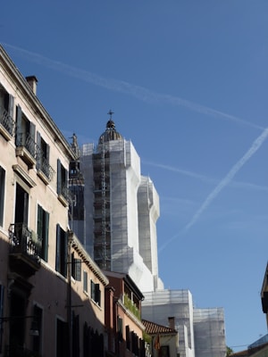 A street view of a historic building under renovation. The structure is wrapped in white scaffolding, with its dome prominently visible against the clear blue sky. The surrounding buildings have traditional architecture with narrow windows and shuttered balconies, adding to the historical ambiance.