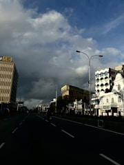 A city street flanked by tall buildings on either side under a dramatic sky with clouds. The road appears empty except for a single motorcycle in the center. Shops and commercial buildings are visible, including a prominent building with an LIC sign.