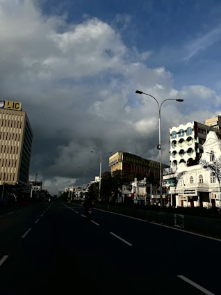A city street flanked by tall buildings on either side under a dramatic sky with clouds. The road appears empty except for a single motorcycle in the center. Shops and commercial buildings are visible, including a prominent building with an LIC sign.