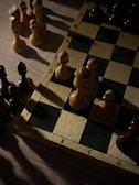 A chessboard is partially visible, featuring several chess pieces arranged on it. The pieces are mostly pawns, with a mix of light and dark colors. The board is set on a wooden surface, and the lighting creates strong contrasts, with shadows cast across the board and pieces.