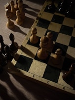 A chessboard is partially visible, featuring several chess pieces arranged on it. The pieces are mostly pawns, with a mix of light and dark colors. The board is set on a wooden surface, and the lighting creates strong contrasts, with shadows cast across the board and pieces.