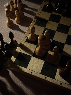 A chessboard is partially visible, featuring several chess pieces arranged on it. The pieces are mostly pawns, with a mix of light and dark colors. The board is set on a wooden surface, and the lighting creates strong contrasts, with shadows cast across the board and pieces.