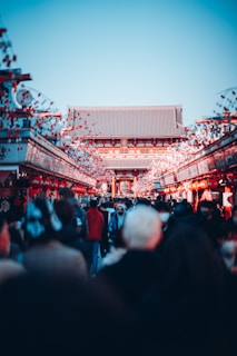 A bustling crowd moves through a vibrant market street lined with decorative red lanterns and banners, leading to a traditional Asian temple with a tiled roof under a clear blue sky.
