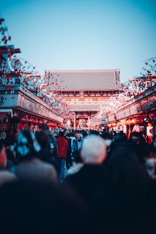 A bustling crowd moves through a vibrant market street lined with decorative red lanterns and banners, leading to a traditional Asian temple with a tiled roof under a clear blue sky.