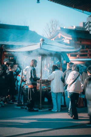 A group of people engages in a traditional ritual around an incense burner in a temple setting, surrounded by structures with traditional architecture. Blue smoke wafts through the air, creating a mystical atmosphere.