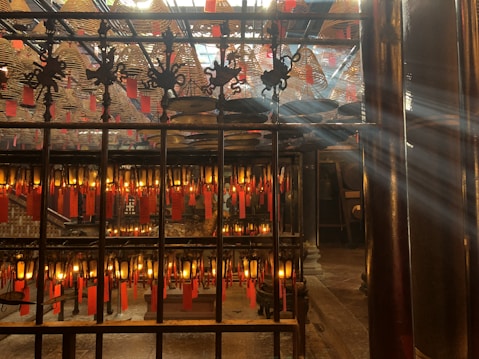 Intricately decorated interior of a traditional temple with numerous hanging spiral incense coils and red prayer tags. Sunlight filters through the space, creating a serene atmosphere. Ornate metalwork and glowing lanterns add to the ambiance.