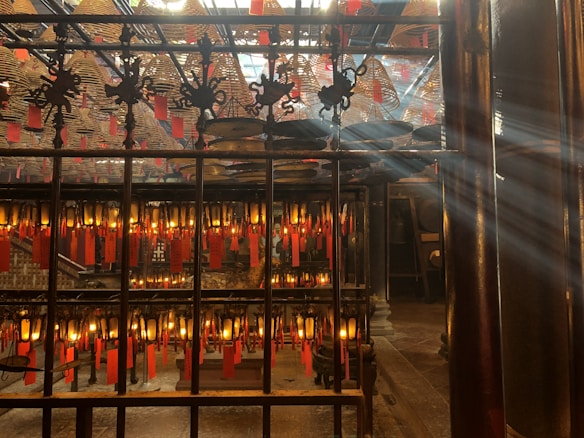 Intricately decorated interior of a traditional temple with numerous hanging spiral incense coils and red prayer tags. Sunlight filters through the space, creating a serene atmosphere. Ornate metalwork and glowing lanterns add to the ambiance.