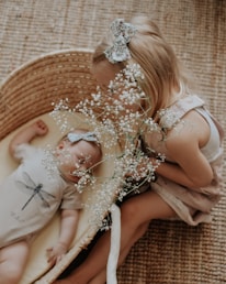 A young child with a bow in their hair is leaning over a wicker basket containing a sleeping baby. Both have matching bows, and the baby is dressed in a onesie with a dragonfly design. The young child holds a branch of delicate white flowers.