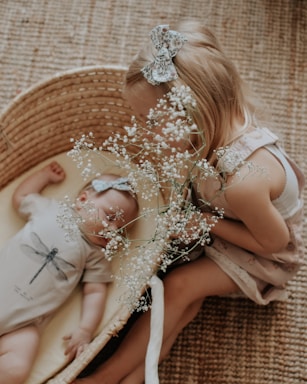 A young child with a bow in their hair is leaning over a wicker basket containing a sleeping baby. Both have matching bows, and the baby is dressed in a onesie with a dragonfly design. The young child holds a branch of delicate white flowers.
