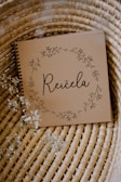 A brown notebook with a spiral binding rests on a woven wicker surface. The cover features delicate floral illustrations forming a circular wreath around the word 'Reičela' in an elegant script. Small white flowers are scattered lightly around, enhancing the rustic and natural aesthetic.