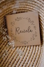 A brown notebook with a spiral binding rests on a woven wicker surface. The cover features delicate floral illustrations forming a circular wreath around the word 'Reičela' in an elegant script. Small white flowers are scattered lightly around, enhancing the rustic and natural aesthetic.