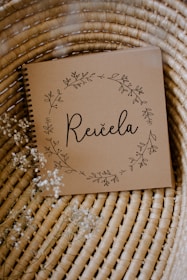 A brown notebook with a spiral binding rests on a woven wicker surface. The cover features delicate floral illustrations forming a circular wreath around the word 'Reičela' in an elegant script. Small white flowers are scattered lightly around, enhancing the rustic and natural aesthetic.