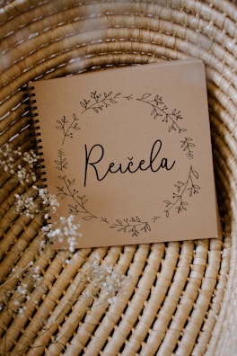 A brown notebook with a spiral binding rests on a woven wicker surface. The cover features delicate floral illustrations forming a circular wreath around the word 'Reičela' in an elegant script. Small white flowers are scattered lightly around, enhancing the rustic and natural aesthetic.