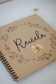A brown kraft paper journal cover features the word 'Reičela' in elegant black script. Delicate floral illustrations adorn the edges, and a pressed daisy is affixed to the cover, adding a natural, handmade touch.